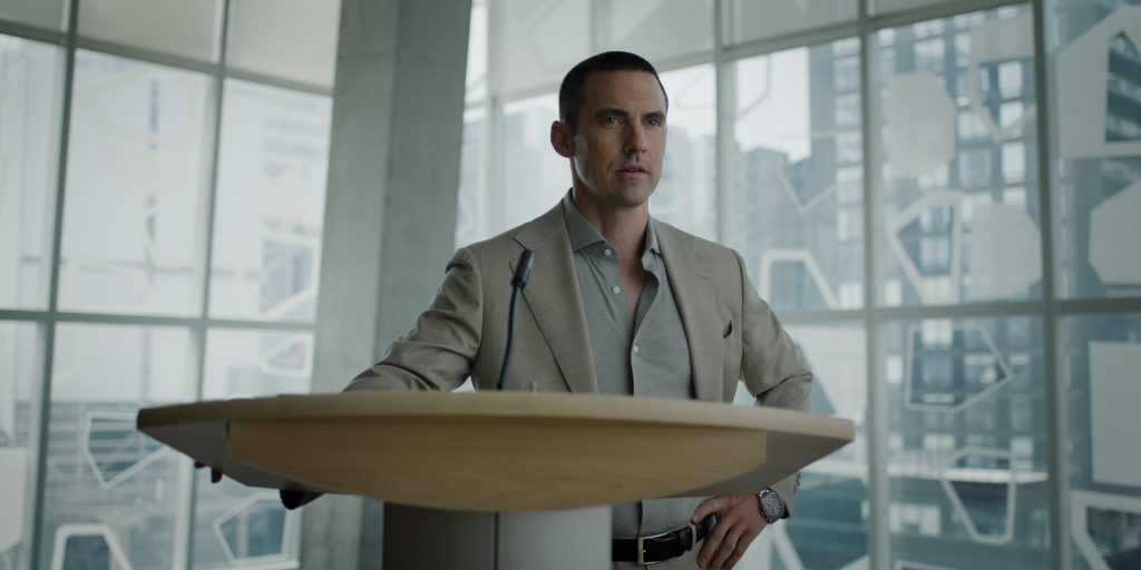 "Man in a suit standing at a podium with a microphone in a modern office or conference room, large windows and cityscape in the background."