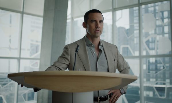 "Man in a suit standing at a podium with a microphone in a modern office or conference room, large windows and cityscape in the background."
